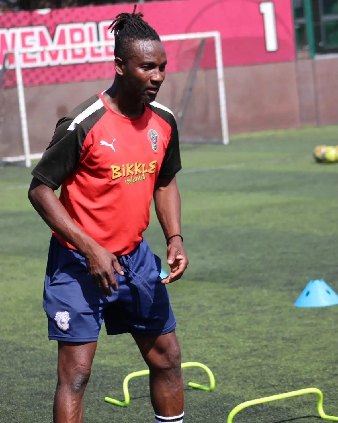 A coach in a red training top on an artificial pitch, with cones and small hurdles laid out for a session