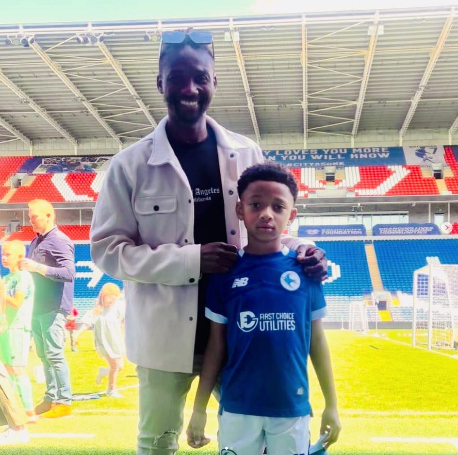 Noah in a blue kit standing beside an adult inside a football stadium