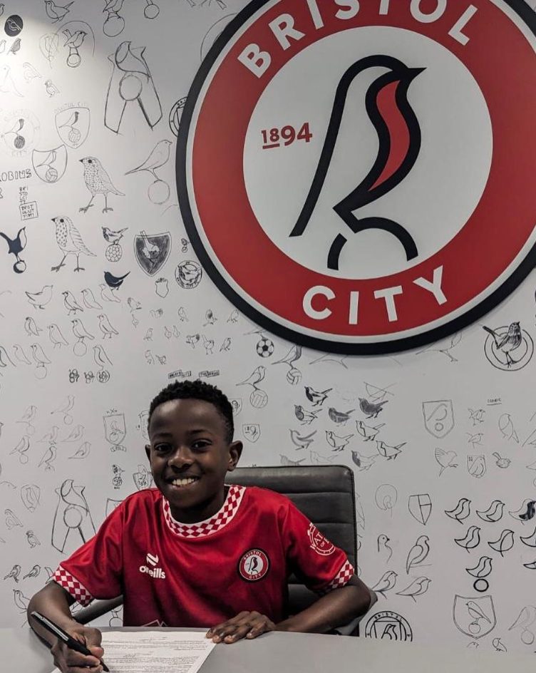 Tiwa smiling at a desk while signing papers, with a club crest on the wall behind