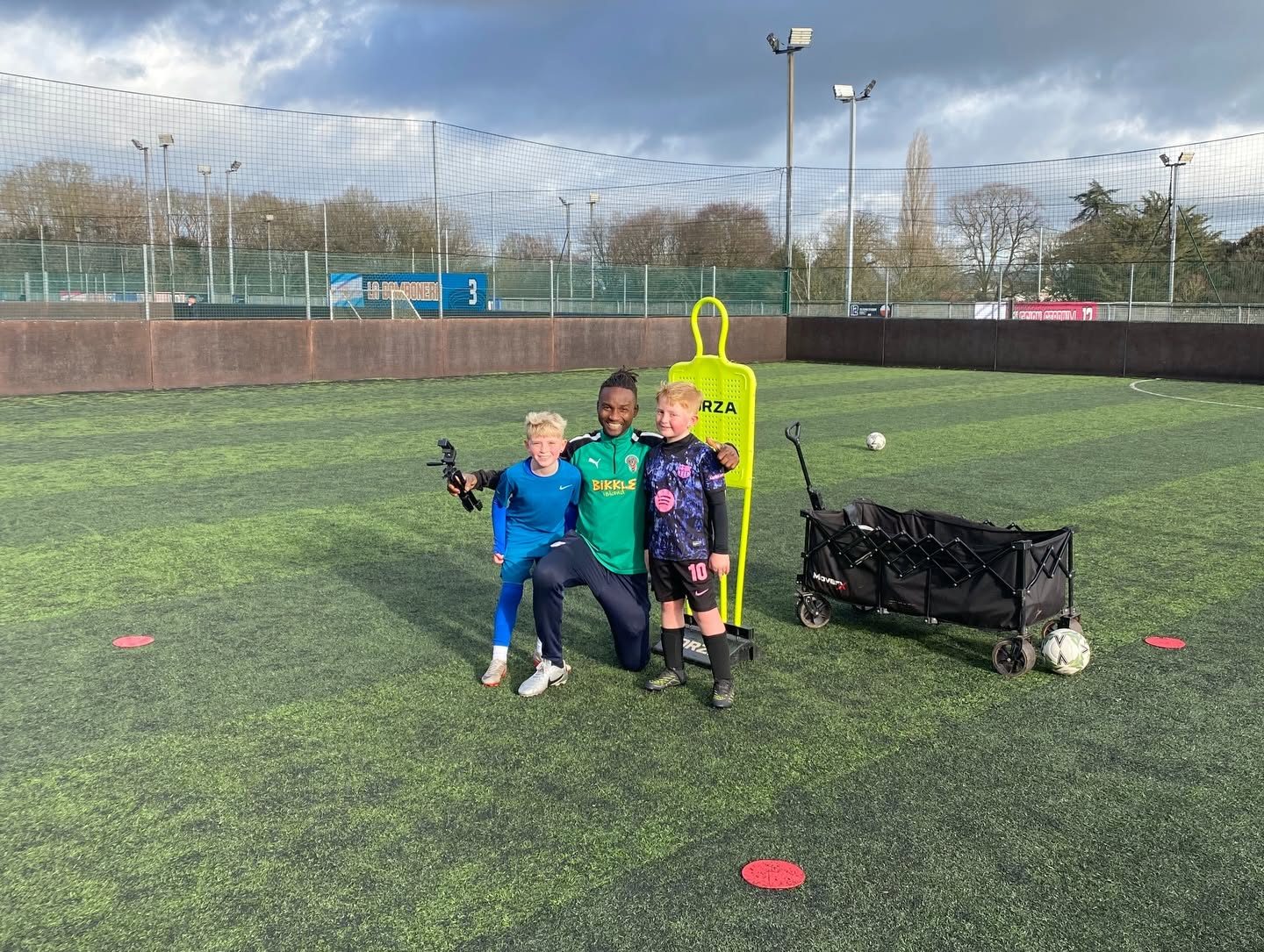 A coach and two young players beside a training mannequin and an equipment trolley on an artificial pitch